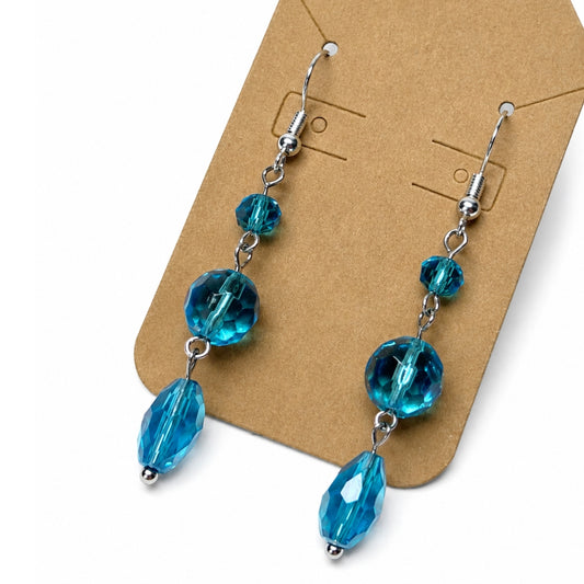 A pair of dangling earrings with faceted blue beads and silver-tone hooks, displayed on a brown kraft card against a white background.