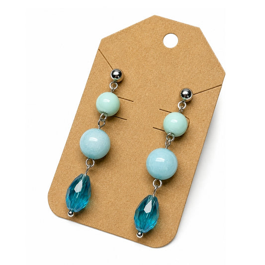 A pair of drop earrings displayed on a brown kraft card against a white background. Each earring has a silver-tone ball stud, a light mint green bead, a larger pale blue bead, and a faceted teal blue teardrop bead.