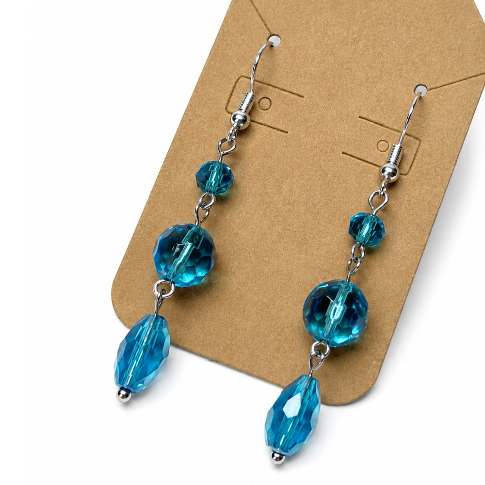 A pair of dangling earrings with faceted blue beads and silver-tone hooks, displayed on a brown kraft card against a white background.